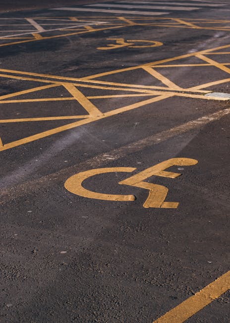 A close-up view of a parking lot detailing designated accessibility spaces marked with bright yellow paint on dark asphalt. The markings include the wheelchair symbol and diagonal lines indicating reserved areas, with visible texture and slight wear on the surface. Surrounding the spaces are neatly painted lines creating clearly defined boundaries, and in the background, additional parking spaces and faded markings can be seen. This image emphasizes organized parking access, relevant to garden and outdoor space management, as highlighted by Gardeners Wimbledon in their guidance on access and parking arrangements for visitors and staff. The lighting suggests a clear day, with shadows cast by the painted markings and the uneven surface texture of the tarmac.