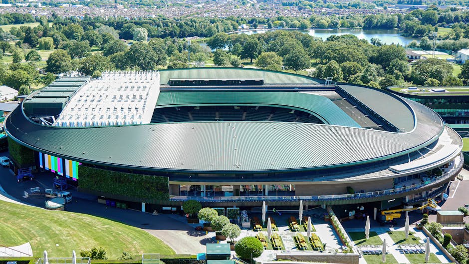 An aerial view of a large, modern, oval-shaped building with a green metal roof, surrounded by well-maintained gardens and pathways. The building features a partially visible open-air section with white slatted roofing and glass-fronted outdoor seating areas, bordered by neatly trimmed hedges and landscaped flower beds with yellow and green plants. Nearby, there's a well-manicured lawn and paved walkways lined with small, rounded shrubs, leading to the entrance area. In the background, lush green trees, a river, and distant countryside under a clear blue sky are visible, creating a peaceful outdoor environment. The scene exemplifies detailed garden maintenance, with attention to landscape layout, plant textures, and outdoor furniture, reflecting the services of Gardeners Wimbledon, especially in relation to planning access and parking for events or visitors.
