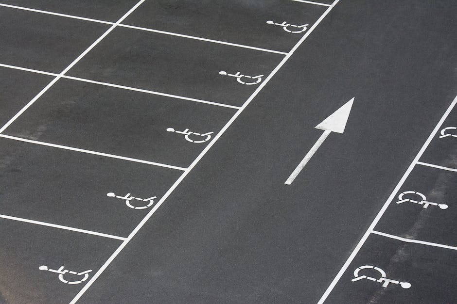 A close-up view of a parking lot detailing designated accessibility spaces marked with bright yellow paint on dark asphalt. The markings include the wheelchair symbol and diagonal lines indicating reserved areas, with visible texture and slight wear on the surface. Surrounding the spaces are neatly painted lines creating clearly defined boundaries, and in the background, additional parking spaces and faded markings can be seen. This image emphasizes organized parking access, relevant to garden and outdoor space management, as highlighted by Gardeners Wimbledon in their guidance on access and parking arrangements for visitors and staff. The lighting suggests a clear day, with shadows cast by the painted markings and the uneven surface texture of the tarmac.