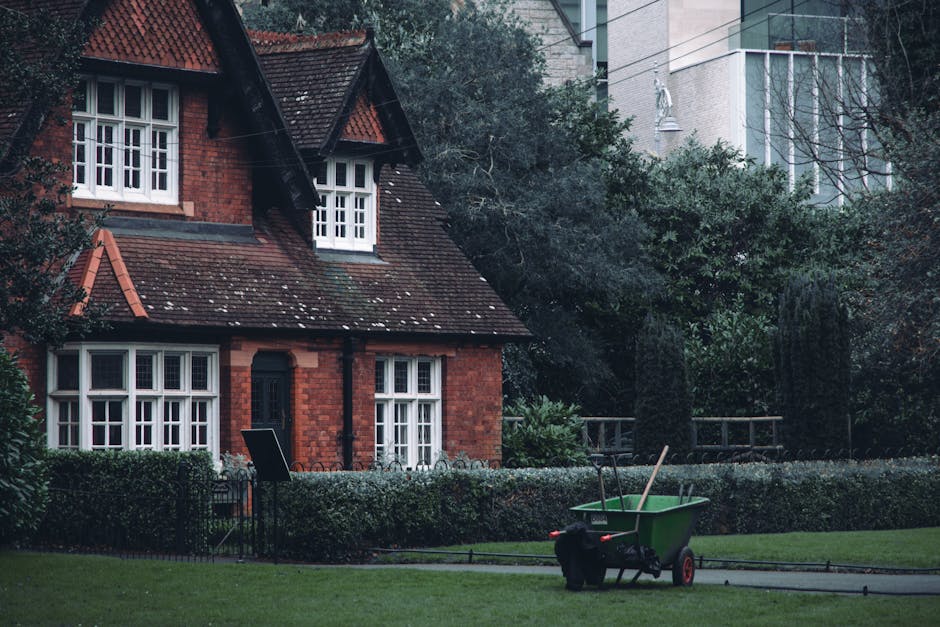 A residential garden scene featuring a traditional red-brick house with a steeply pitched roof and white-framed windows, surrounded by well-maintained hedges and lush trees. In the foreground, there is a neatly mowed lawn with visible grass textures and a pathway. A green garden wheelbarrow, filled with gardening tools such as a pitchfork and a rake, stands on the grass, indicating ongoing gardening or lawn care activities. The background shows a mix of mature trees and shrubbery, with some tall modern buildings subtly visible beyond the foliage, reflecting a blend of urban and suburban environments. The lighting is natural, suggesting an overcast day suitable for outdoor maintenance. The scene conveys a professional gardening setting, consistent with the services offered by Gardeners Wimbledon in garden maintenance and landscaping work.