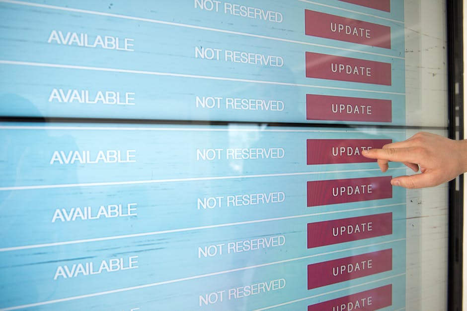 A person pointing at a digital screen displaying appointment slots labeled 'AVAILABLE' and 'NOT RESERVED' with red 'UPDATE' buttons next to the available slots. The screen is mounted outdoors, with the reflection of buildings and sky visible on its surface. The scene suggests scheduling or managing garden maintenance appointments, emphasizing the process of booking landscaping work or lawn care services. The environment appears bright with natural lighting, though no specific garden elements such as plants or tools are visible in this image.