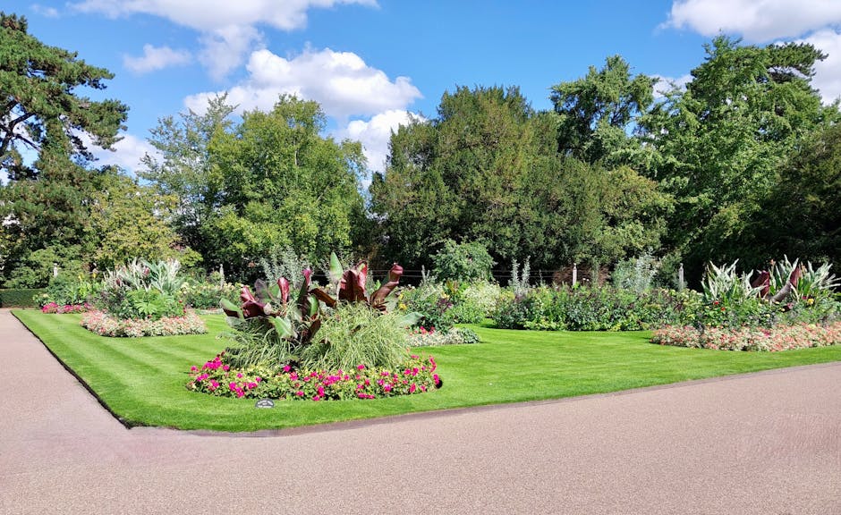 A residential garden scene featuring a traditional red-brick house with a steeply pitched roof and white-framed windows, surrounded by well-maintained hedges and lush trees. In the foreground, there is a neatly mowed lawn with visible grass textures and a pathway. A green garden wheelbarrow, filled with gardening tools such as a pitchfork and a rake, stands on the grass, indicating ongoing gardening or lawn care activities. The background shows a mix of mature trees and shrubbery, with some tall modern buildings subtly visible beyond the foliage, reflecting a blend of urban and suburban environments. The lighting is natural, suggesting an overcast day suitable for outdoor maintenance. The scene conveys a professional gardening setting, consistent with the services offered by Gardeners Wimbledon in garden maintenance and landscaping work.